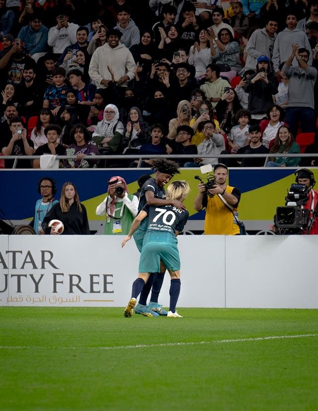 Two players embrace in celebration while a press photographer and a broadcast cameraman film them pitchside at Match for Hope in Doha