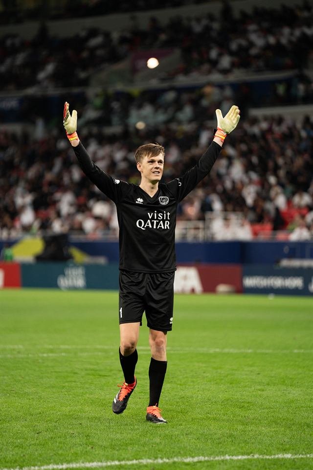 A goalkeeper in a black Visit Qatar kit walks across the pitch with both arms raised to the crowd at Match for Hope in Doha